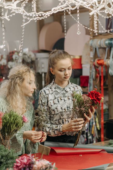 A young woman and an older woman are standing at a floral arrangement station in a floral shop, both holding small bouquets of fresh flowers. The young woman is holding a collection of red roses and g