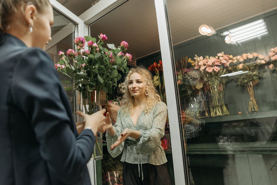 A woman with curly blonde hair and a patterned blouse receives a bouquet of vibrant pink roses from a florist inside a shop, with shelves of various flower arrangements in glass vases visible in the b