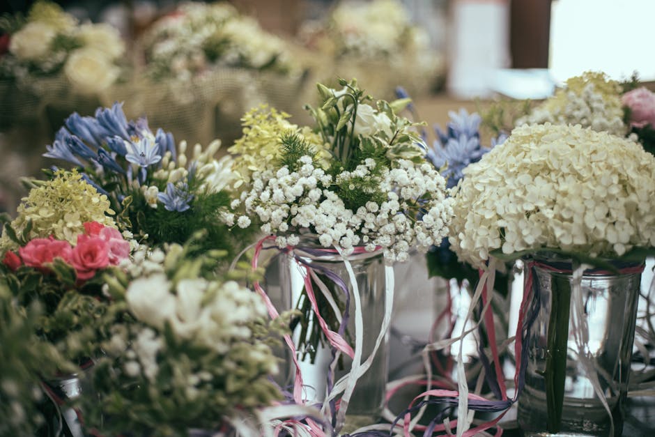 An arrangement of freshly cut flowers displayed in clear glass vases, featuring white hydrangeas, light pink roses, sprigs of blue agapanthus, and delicate white baby's breath. The flowers are arrange