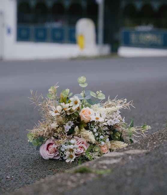 A delicate bouquet of fresh flowers resting on an asphalt pavement near the curb, featuring soft pink roses, white daisies, and small white filler flowers, complemented by greenery and wispy dried ele