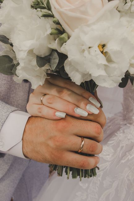Close-up of a bride and groom holding a floral bouquet during a wedding ceremony. The bride's hand, adorned with a silver wedding band and decorated nails, gently wraps around the groom's hand, which 