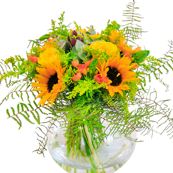 The spirit of Barkingside in high summer is captured within this radiant bouquet, where bold yellow sunflowers and plush garden roses come together in a statement of pure happiness. Large sunflower heads beam outward, their golden petals and dark mahogany centers commanding attention, while velvet-petaled roses curl around them in gentle, embracing spirals. Between these stars, bright red hypericum berries glint like scattered rubies, enhancing the color contrast and adding a touch of festive cheer. Ferns and delicate sprays of green foliage arch and cascade over the vase's rim, reminiscent of the abundant greenery that lines the paths of Barkingside Recreation Ground. Pale yellow filler flowers - perhaps local solidago - and elegant eucalyptus lend lightness and texture, their hues mingling for a transparent, multi-layered finish. All are anchored by the clean lines of the crystal sphere vase, where every vibrant stem stands in water, testament to the arrangement's freshness and care. The subtle fragrance of sun-kissed blooms and cut greenery lingers, softly infusing the room with the promise of good days. Ideal as a focal piece for milestone events across Barkingside or a bright gesture delivered to a friend near Gants Hill, this bouquet is a true expression of local artistry and seasonal abundance.